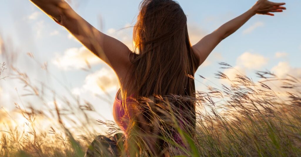 Жена Седи в Поле с Гръб и Разперени Ръце Като Птица Към Небето. Носи Лилав Потник и Гледа Към Слънцето Пред Нея.